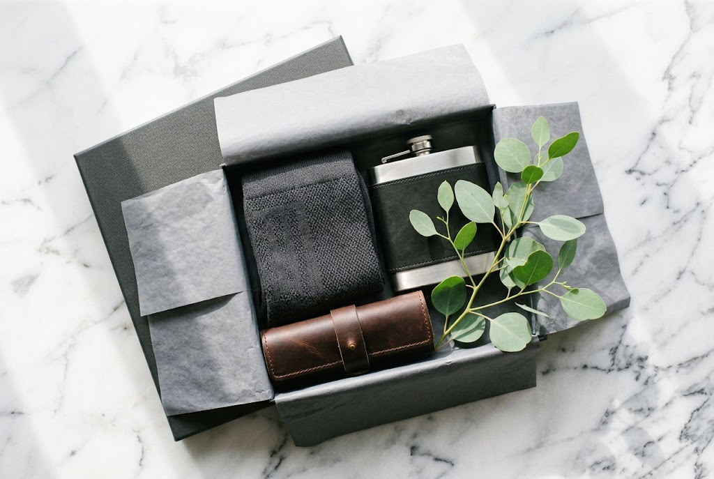 Open gift box with folded charcoal socks, a hip flask and eucalyptus on marble.